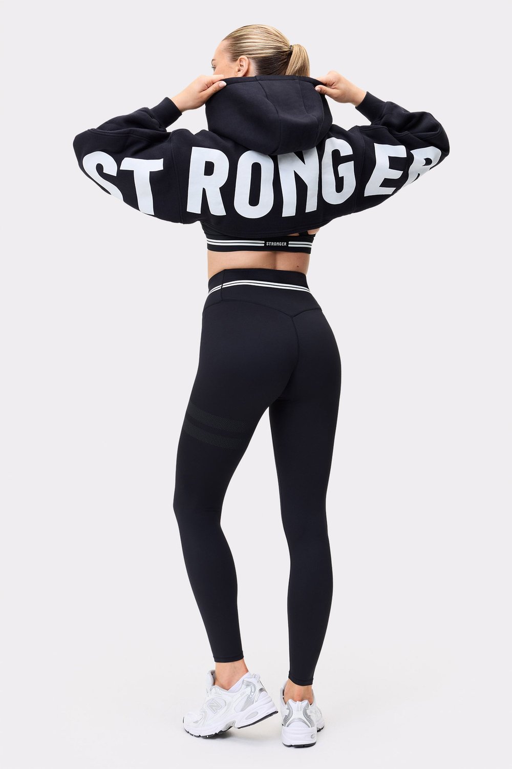Rückansicht eines Models im schwarzen Cropped Hoodie von Stronger mit auffälligem weißen Schriftzug auf den Ärmeln.