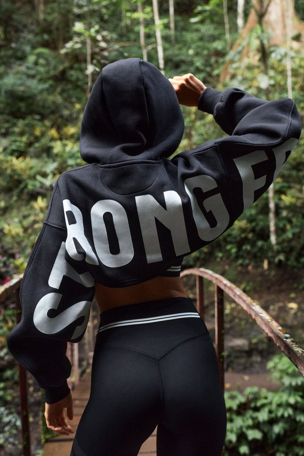 Rückansicht eines Models in dem schwarzen Cropped Hoodie von Stronger mit großem Logodesign auf den Ärmeln.