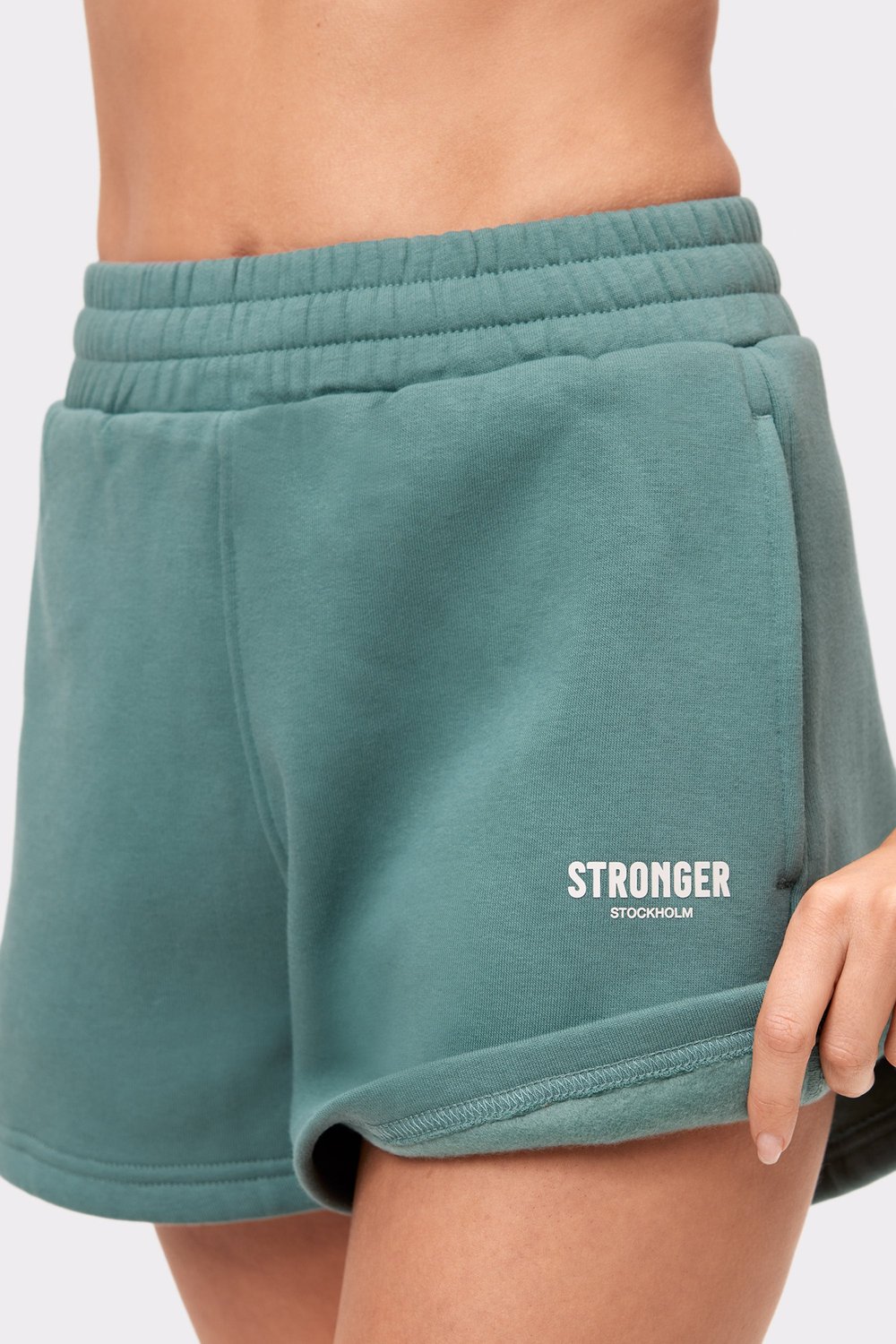 Nahaufnahme der grünen Comfy Sweatshorts von Stronger mit weißem „Stronger Stockholm“-Logo auf dem linken Oberschenkel.