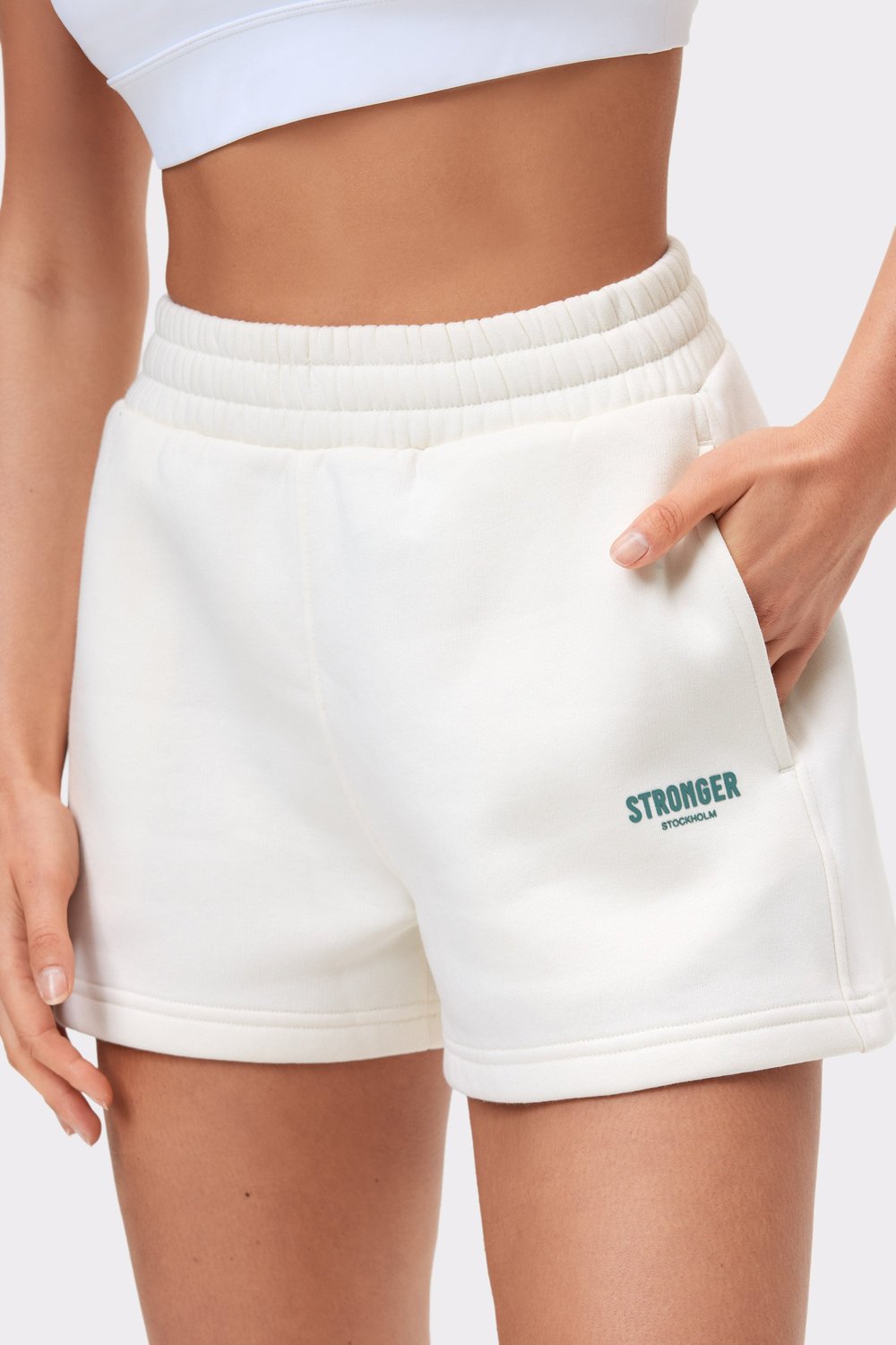 Nahaufnahme der weißen Comfy Sweatshorts von Stronger, die den hohen elastischen Bund, die Seitentasche und das Logo betont.