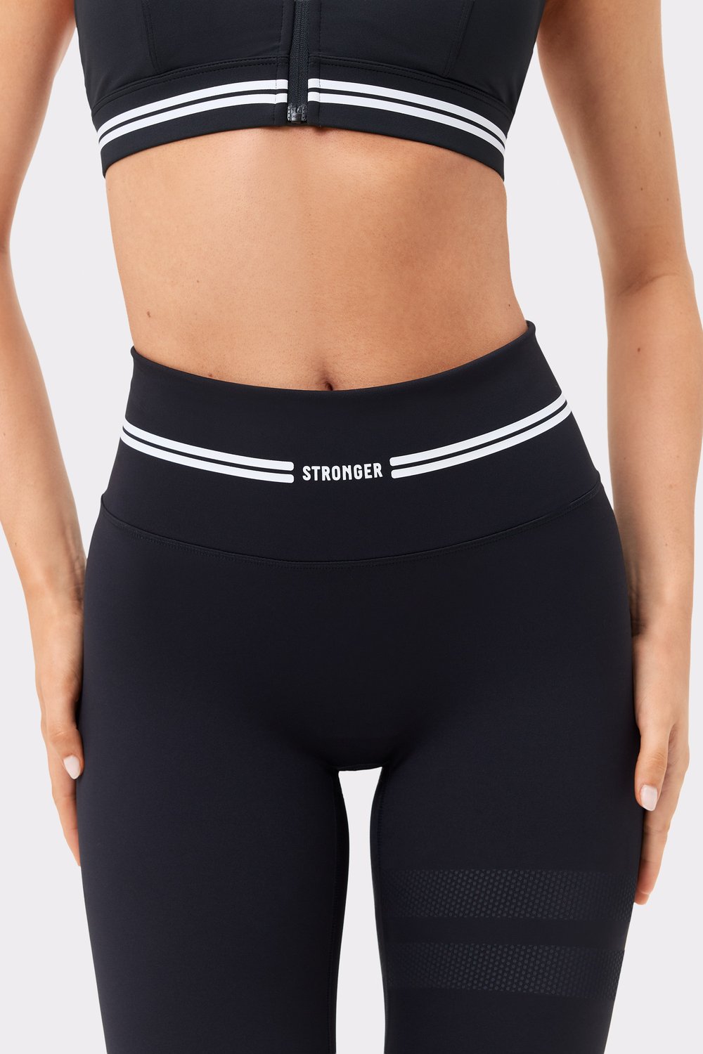 Close-up on the high waist of black Signature Logo Leggings, featuring white signature stripes and the Stronger logo.