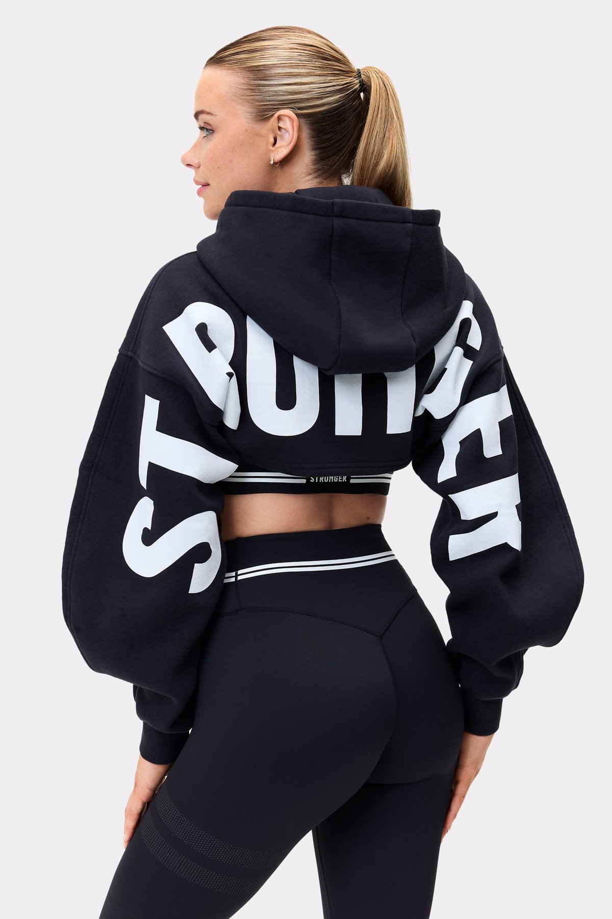 Model trägt den schwarzen Cropped Hoodie von Stronger mit auffälligem weißen Ärmelschriftzug und Oversized-Passform.