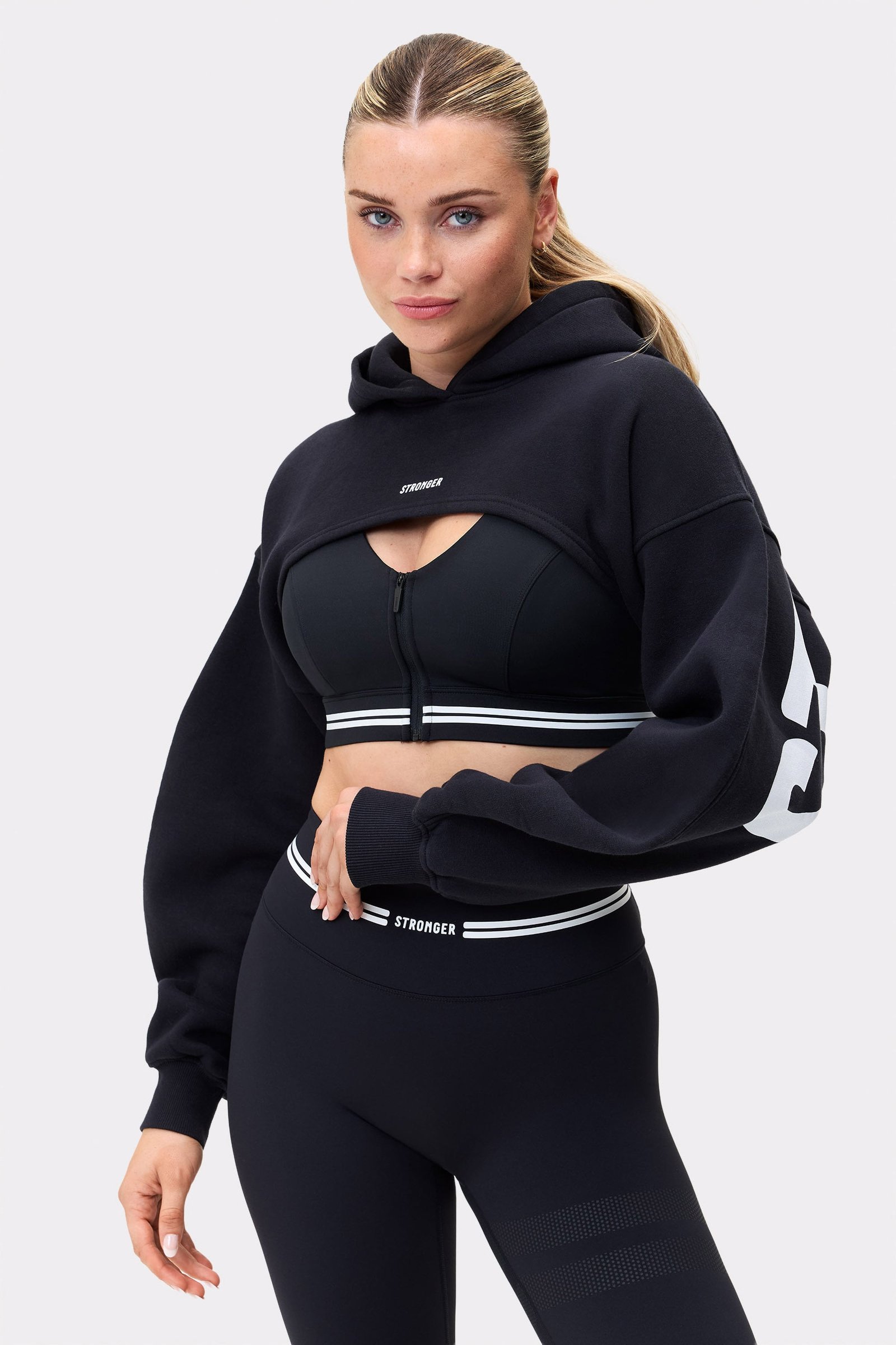 Model trägt den schwarzen Cropped Hoodie von Stronger, ein Oversized-Oberteil mit abgerundeter Vorderseite und Logodetails.