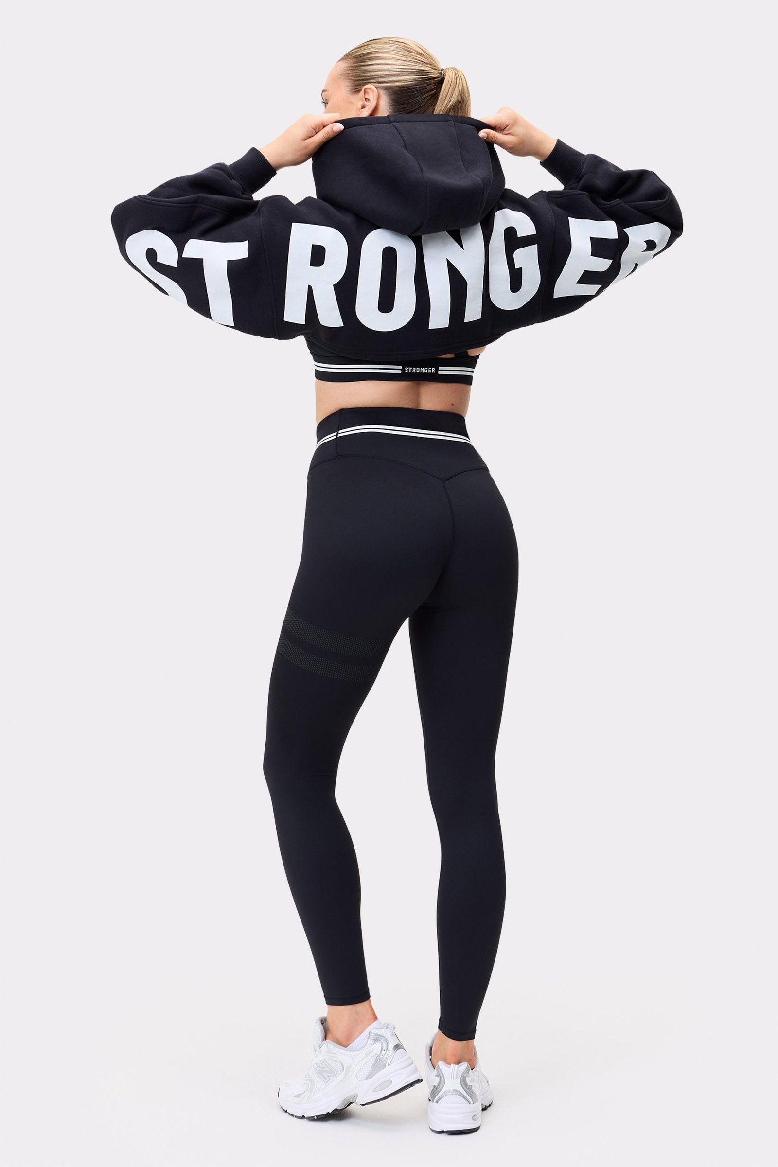 Rückansicht eines Models im schwarzen Cropped Hoodie von Stronger mit auffälligem weißen Schriftzug auf den Ärmeln.