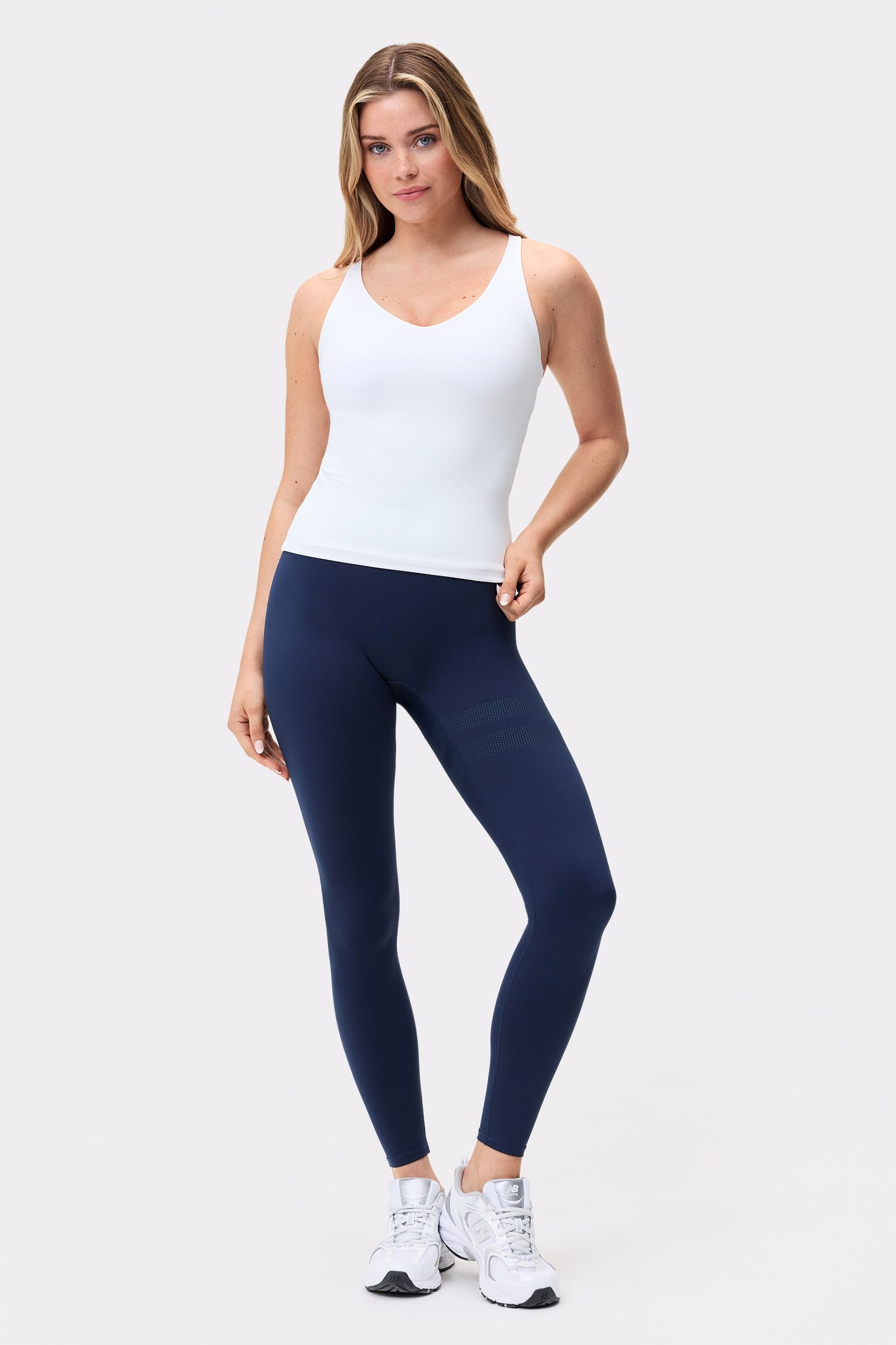 Model trägt das weiße Signature Strappy Tank von Stronger, ein figurbetontes Oberteil mit dunklen Leggings.