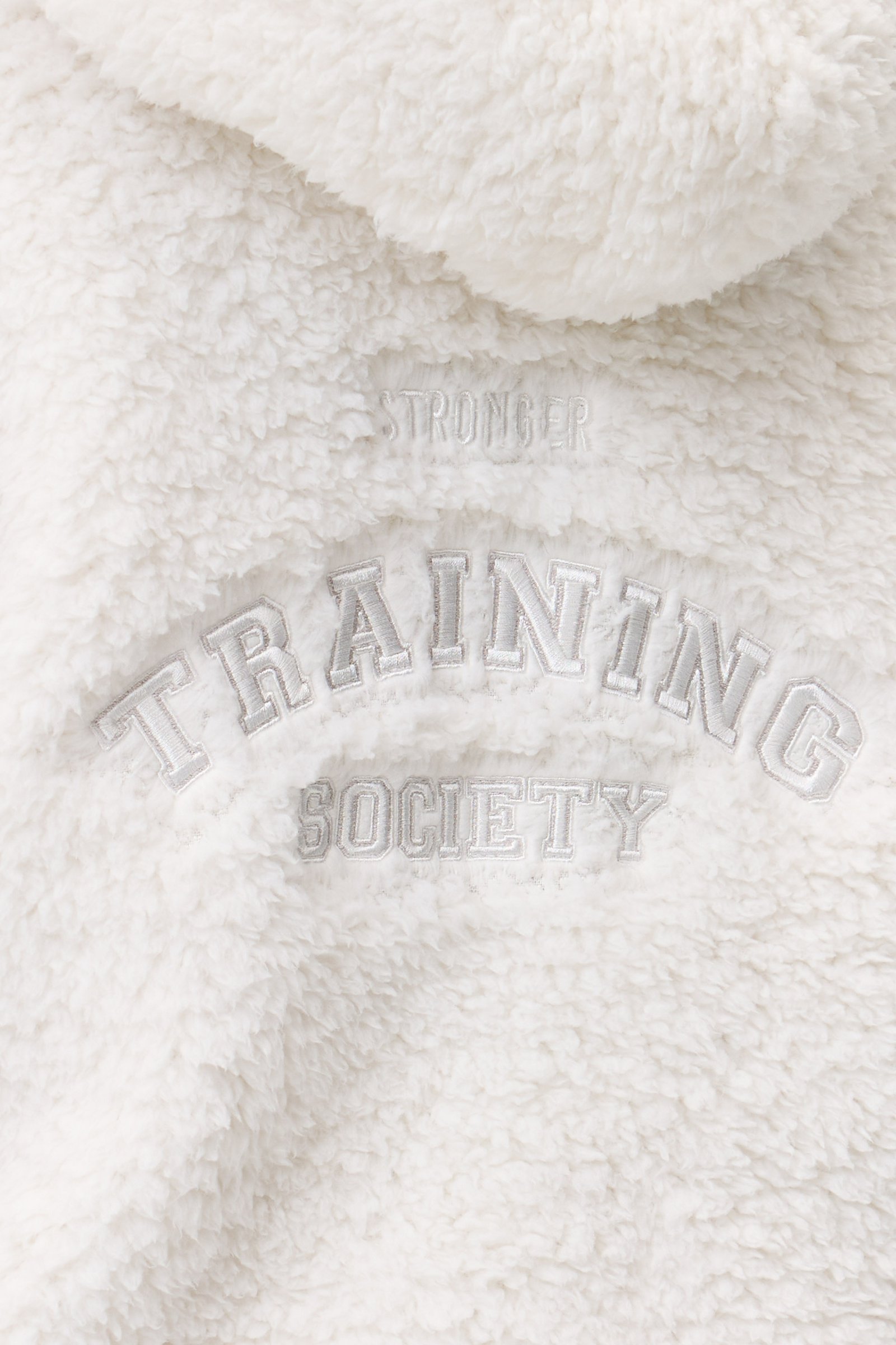 Nahaufnahme des Logos auf der weißen Hooded Pile Jacket von Stronger mit weicher Textur aus recyceltem Polyester.