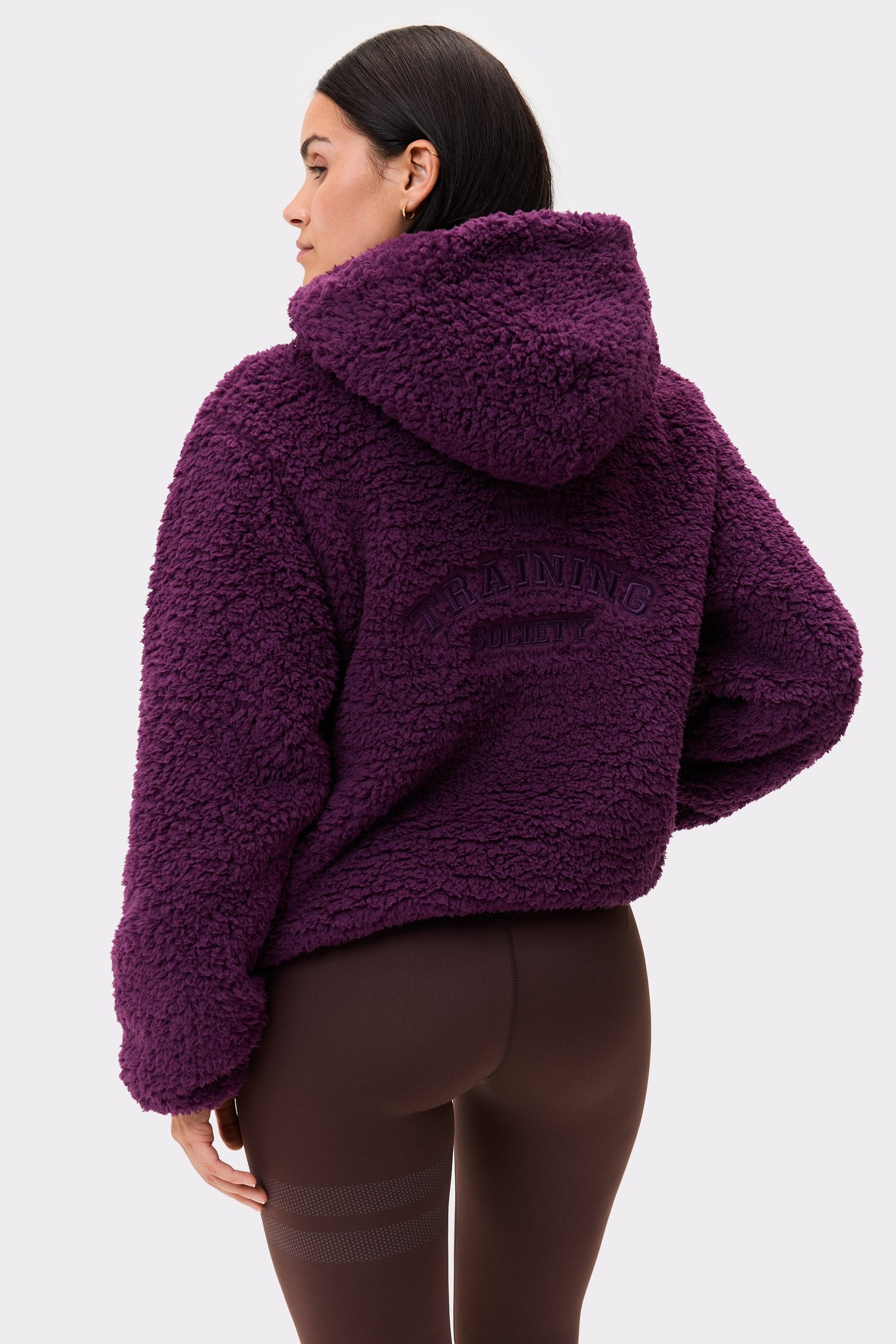 Back view of a model in the dark purple Hooded Pile Jacket from stronger, showing its texture, hood, and back embroidery.