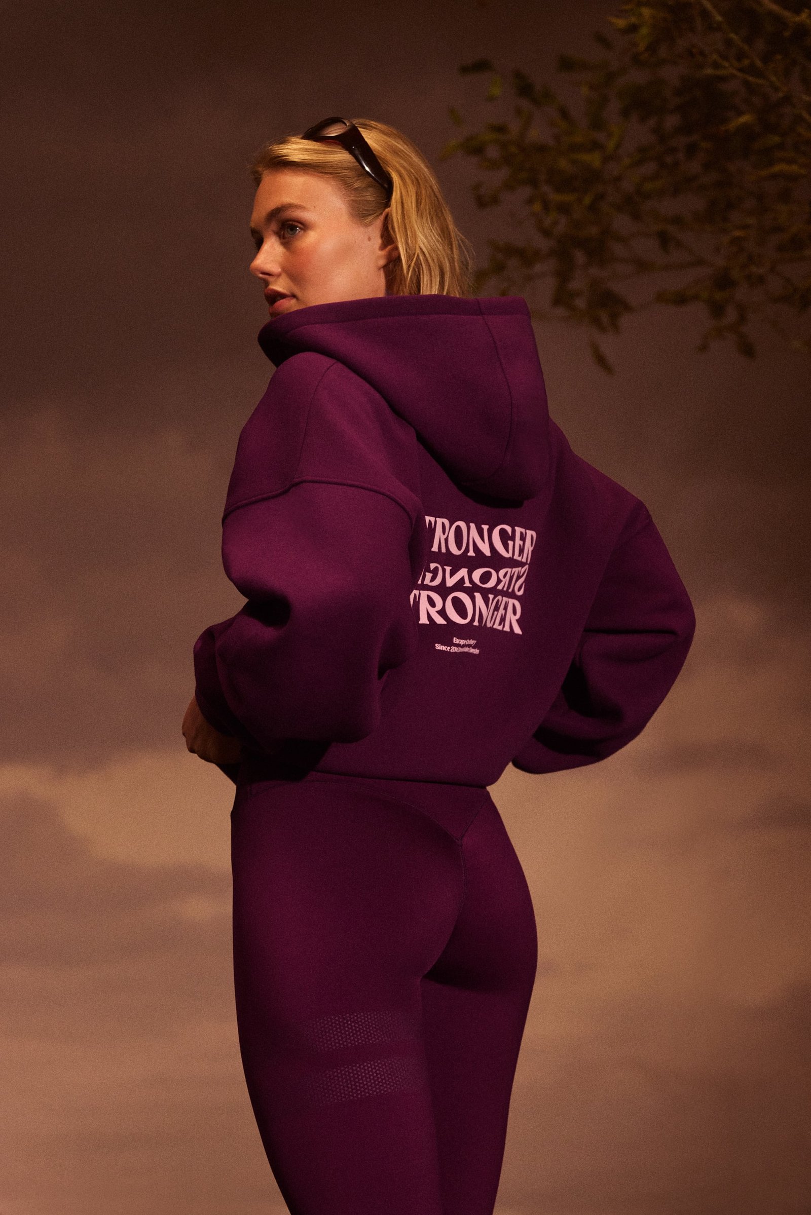 Back view of a model wearing the Relaxed Full Zip Hoodie in aubergine purple from Stronger, showing the back logo.